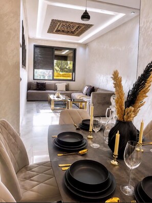 A modern dining area with a long table set for four, featuring black plates, gold cutlery, and candles.