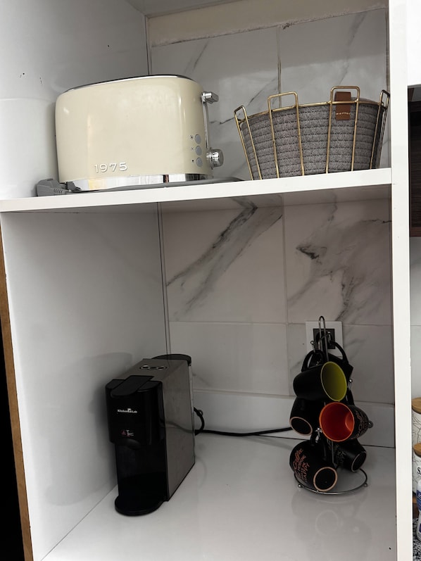 A kitchen with a white countertop, a toaster, a coffee maker, and a shelf with a basket and a kettle.