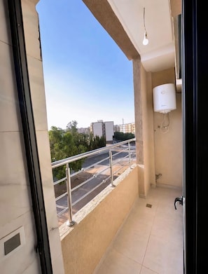 A balcony with a metal railing, a water heater mounted on the wall, and a clear view of a road and buildings in the distance.