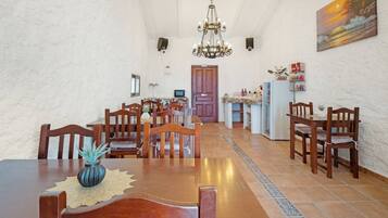 A dining area with wooden tables and chairs, a chandelier, and a kitchen in the background.