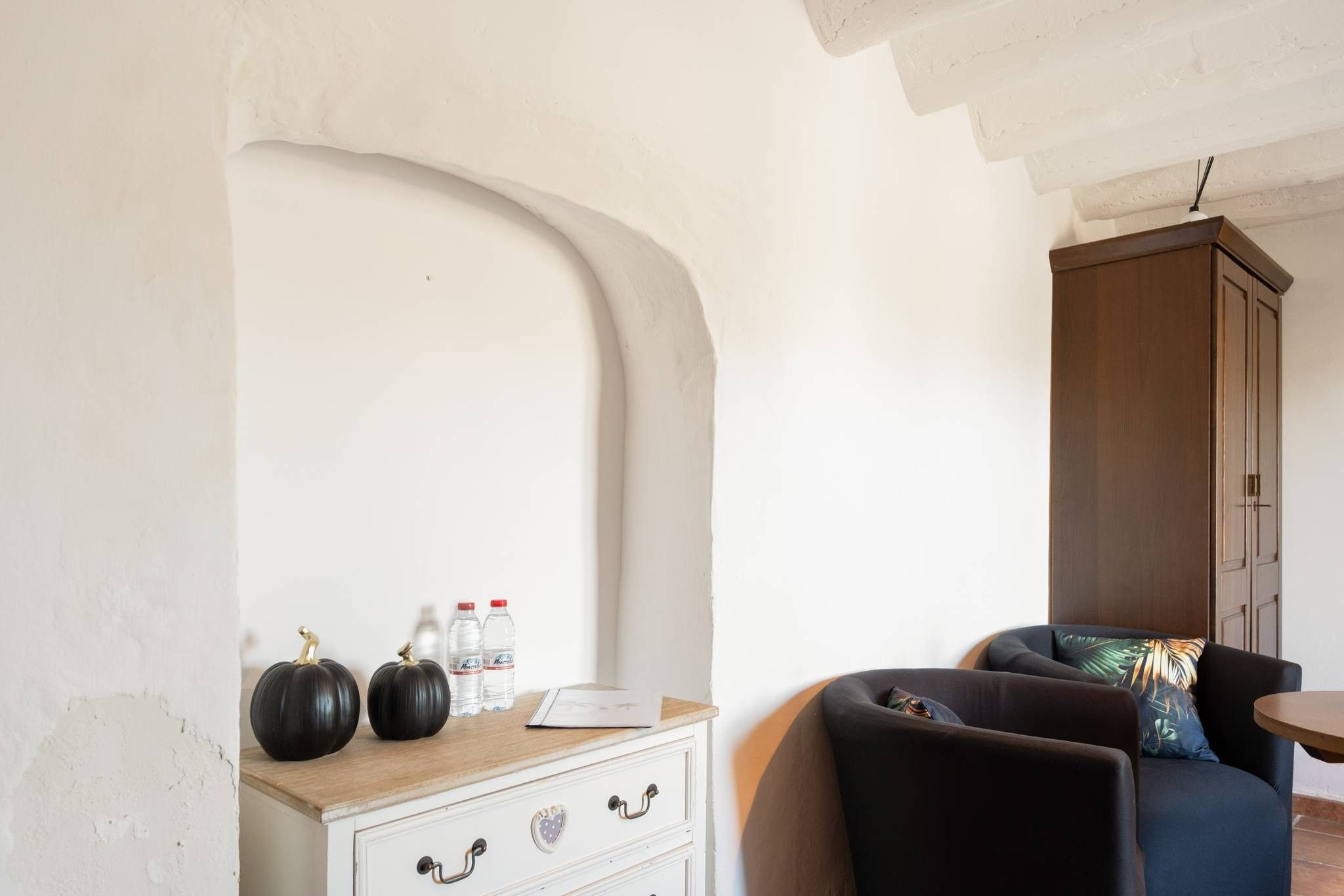 Una habitación con un arco blanco, un aparador de madera con calabazas decorativas, un sofá oscuro con un cojín tropical y un mueble de madera.