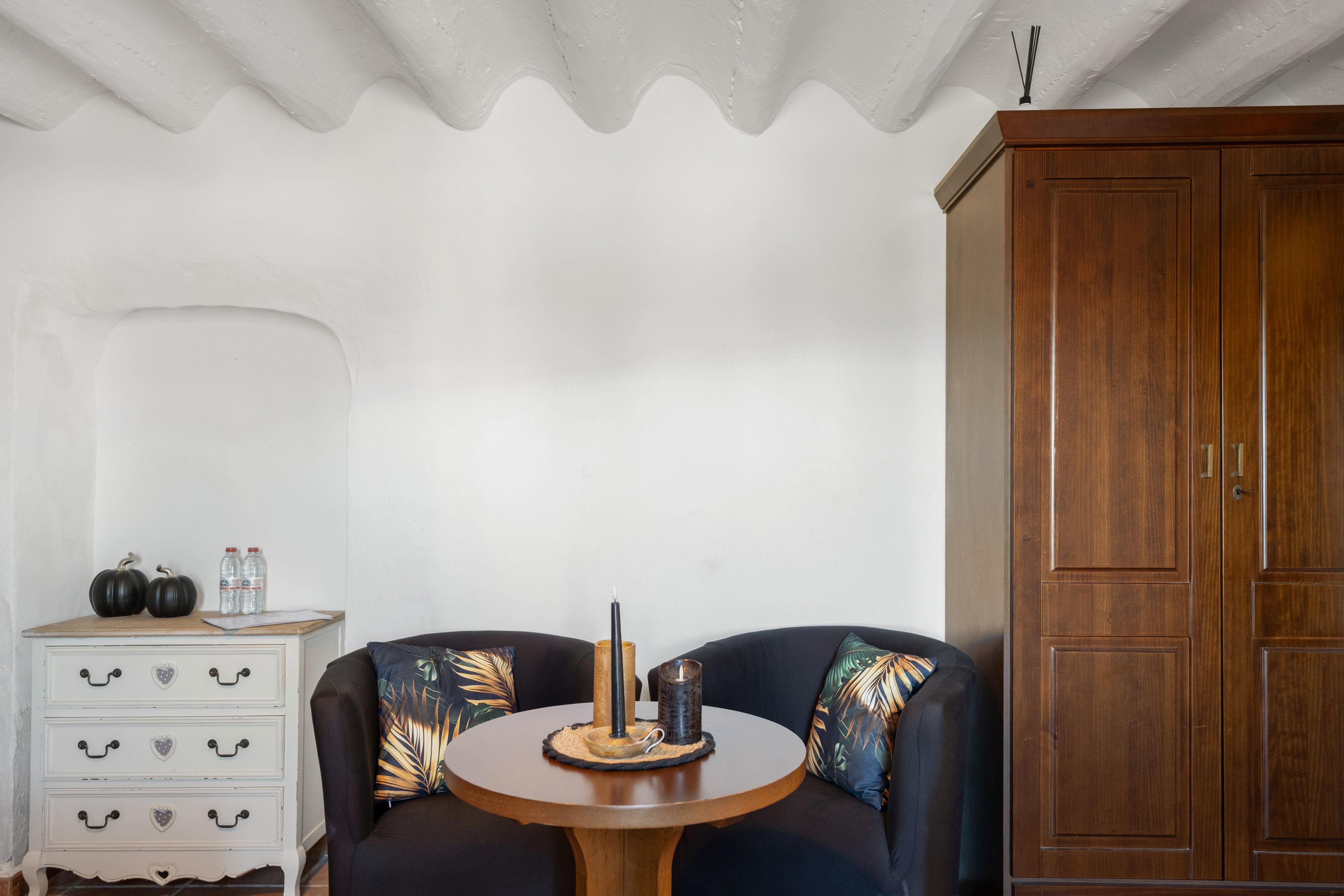 A room with a round wooden table, two black sofas with tropical leaf pillows, a white dresser with drawers, and a wooden wardrobe.