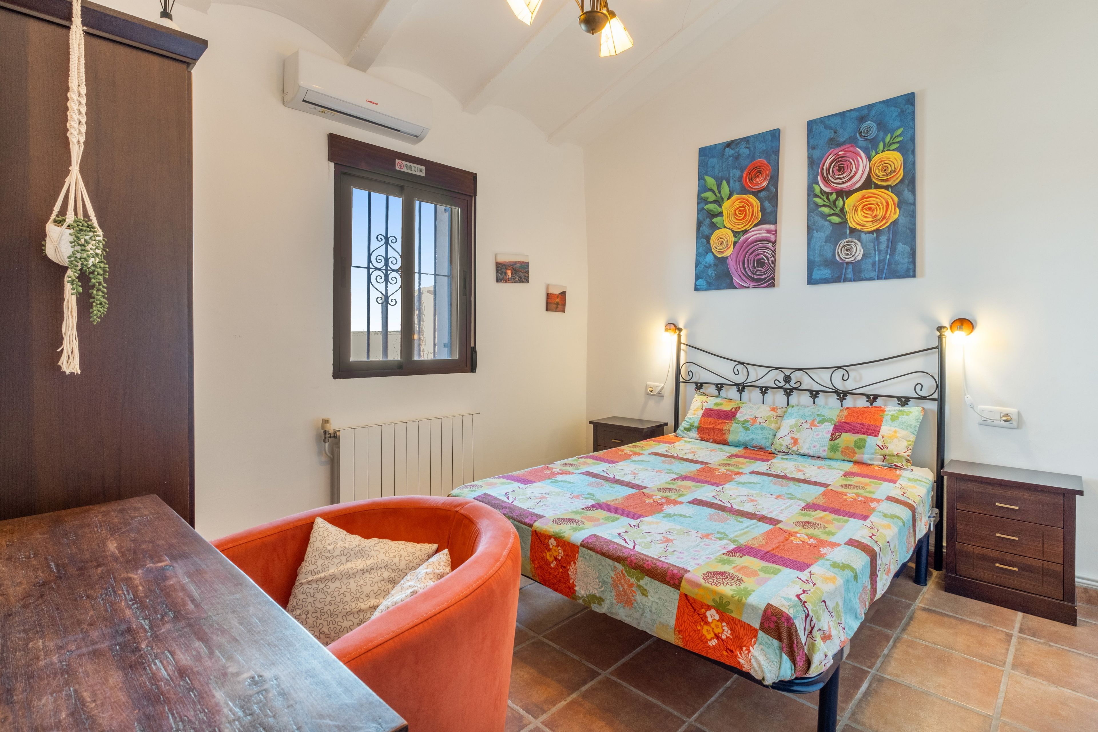 Un dormitorio con cama, una silla, mesita de noche, cómoda, radiador, ventana con rejas y dos cuadros colgados en la pared.