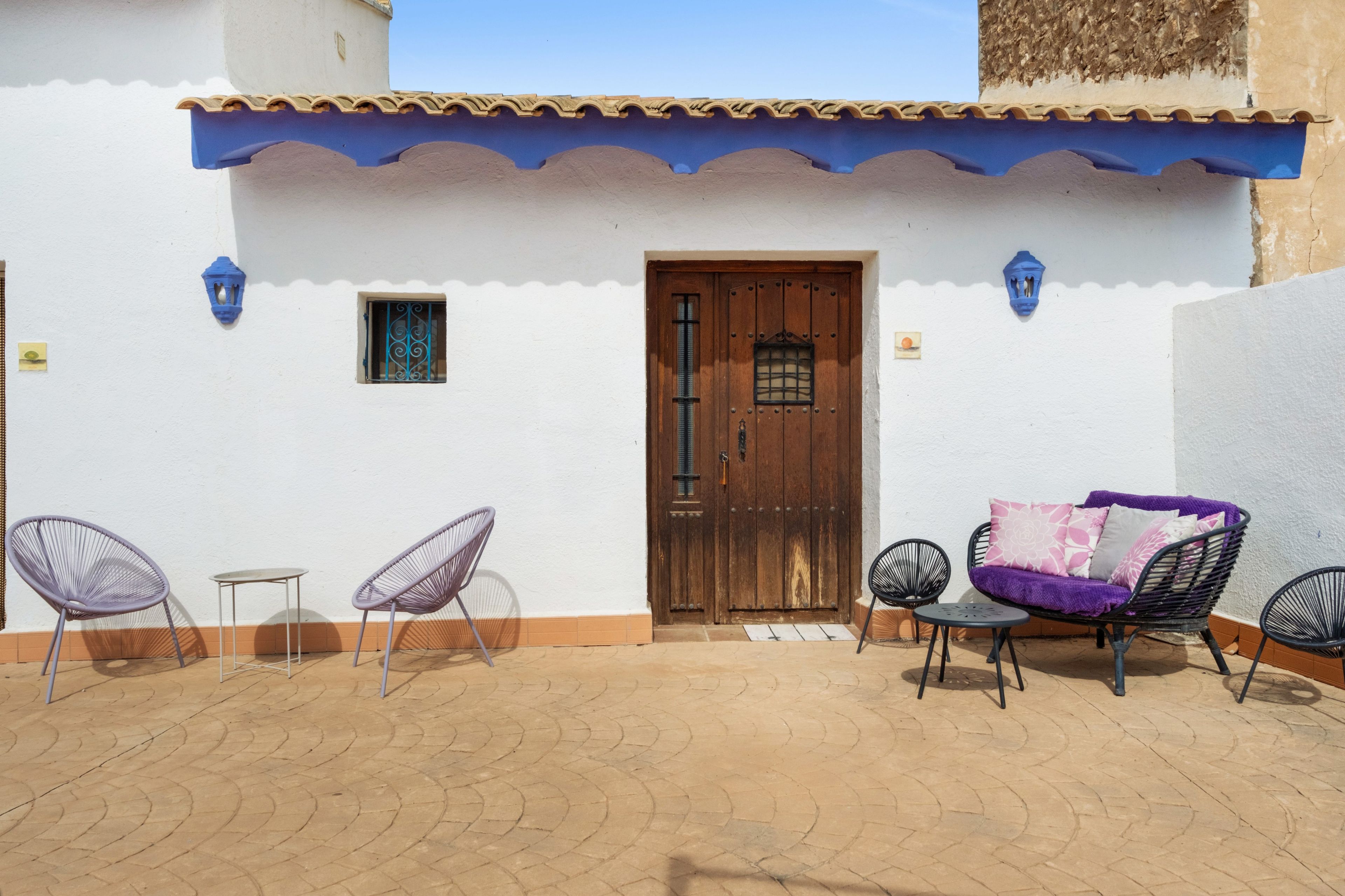 Un patio con un edificio blanco, una puerta de madera, dos sillas, una mesita y un sofá con cojines.