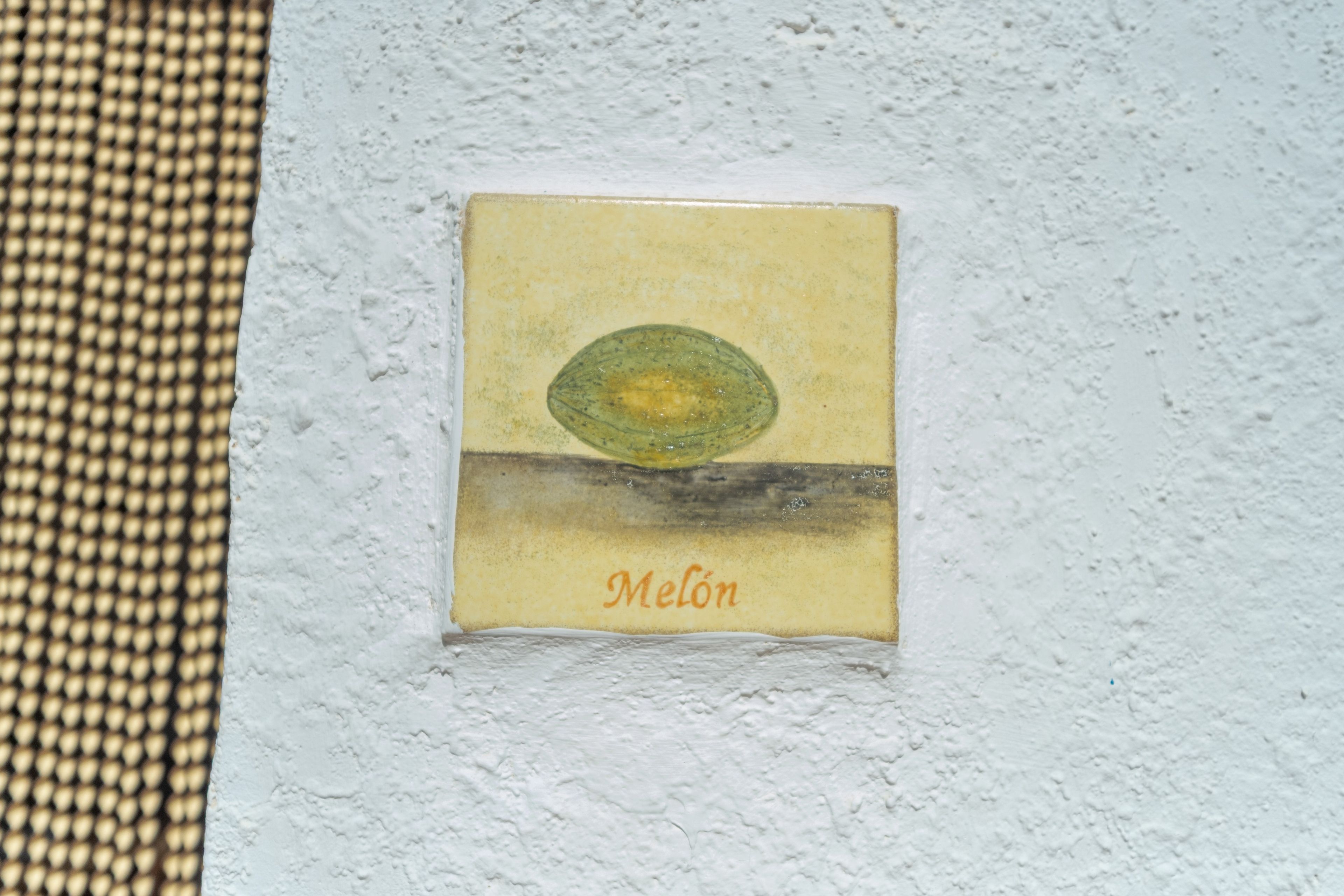 Un objeto pequeño y cuadrado, montado en la pared, con textura y un diseño ovalado en el centro, y la palabra 'Melón' en la parte inferior.