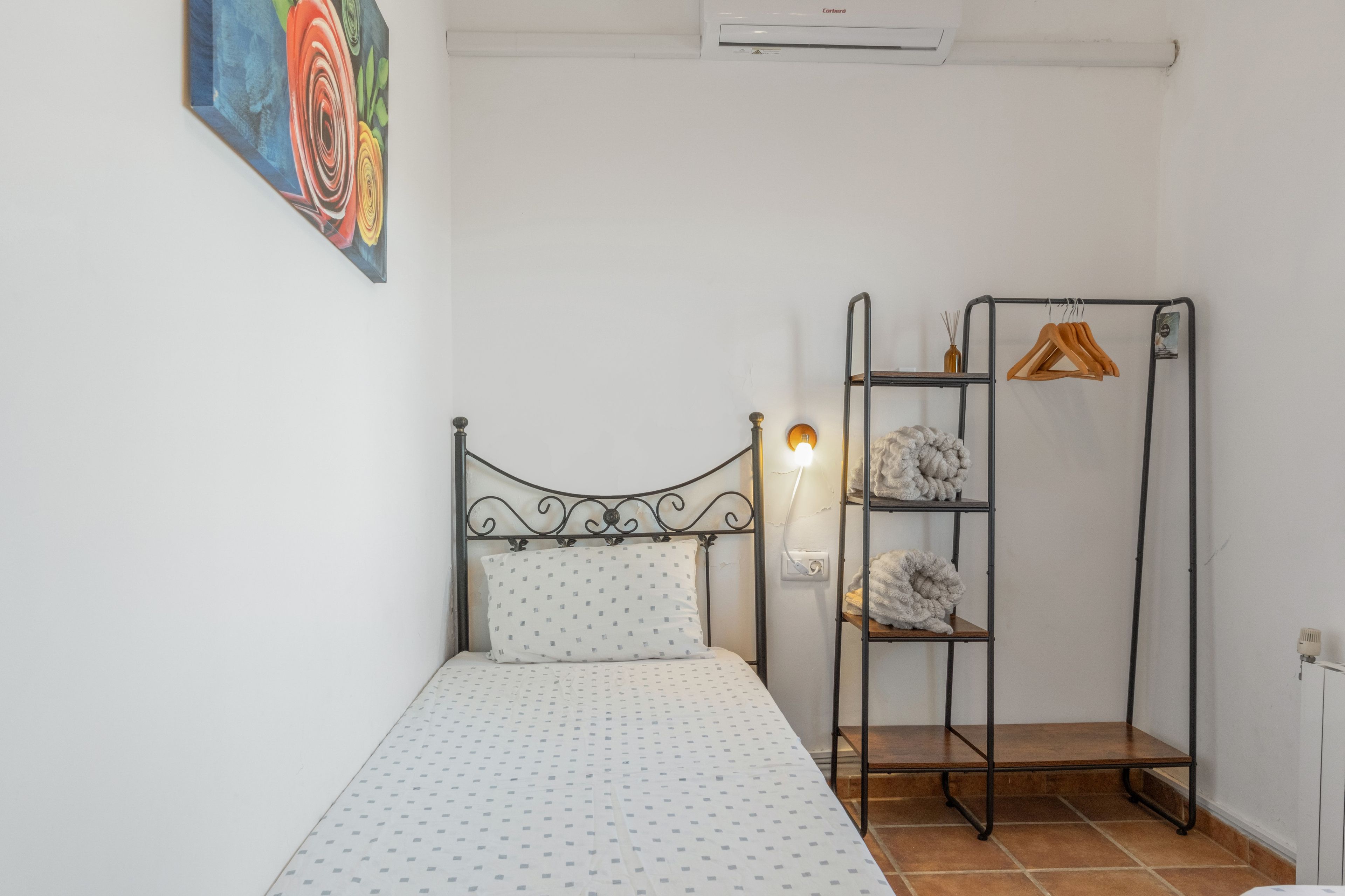 Un dormitorio pequeño y ordenado con una cama individual, una estantería metálica con ropa colgada y un cuadro colorido en la pared.