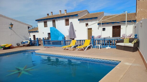 Une piscine avec des chaises longues, un mur bleu et un bâtiment doté d’un toit en tuiles.