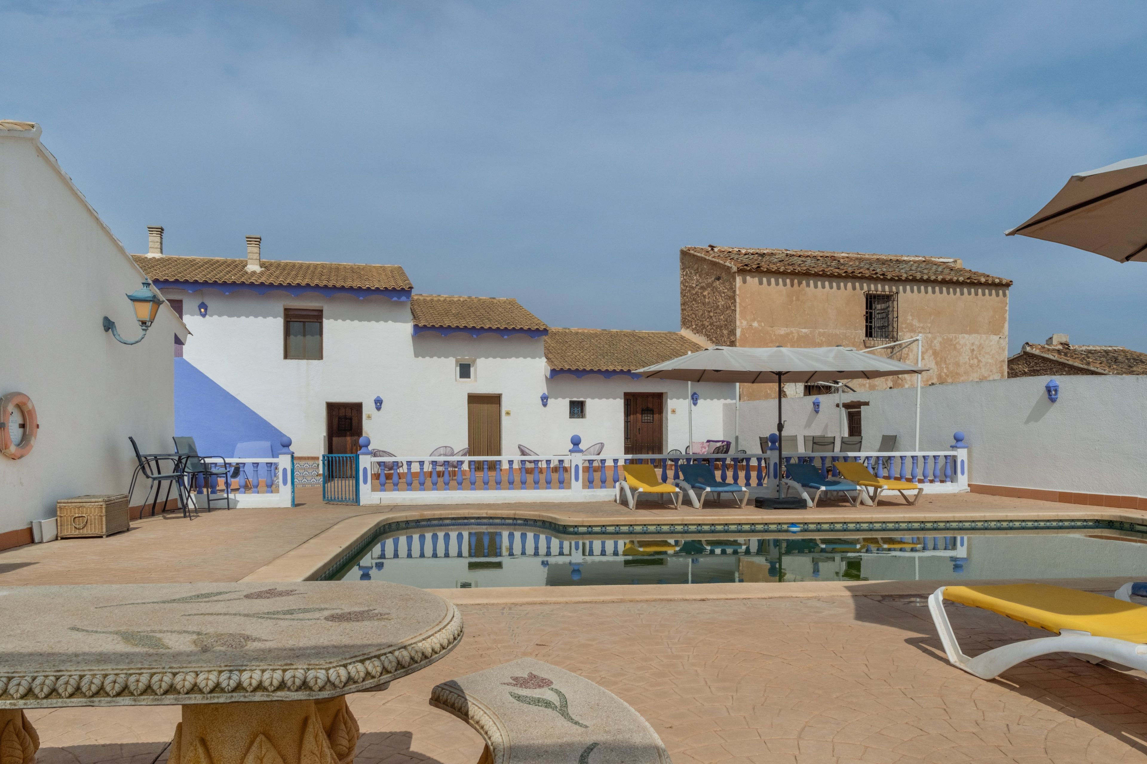 A pool area with lounge chairs, a table, and a building with a tiled roof.