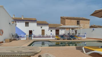 A pool area with lounge chairs, a table, and a building with a tiled roof.