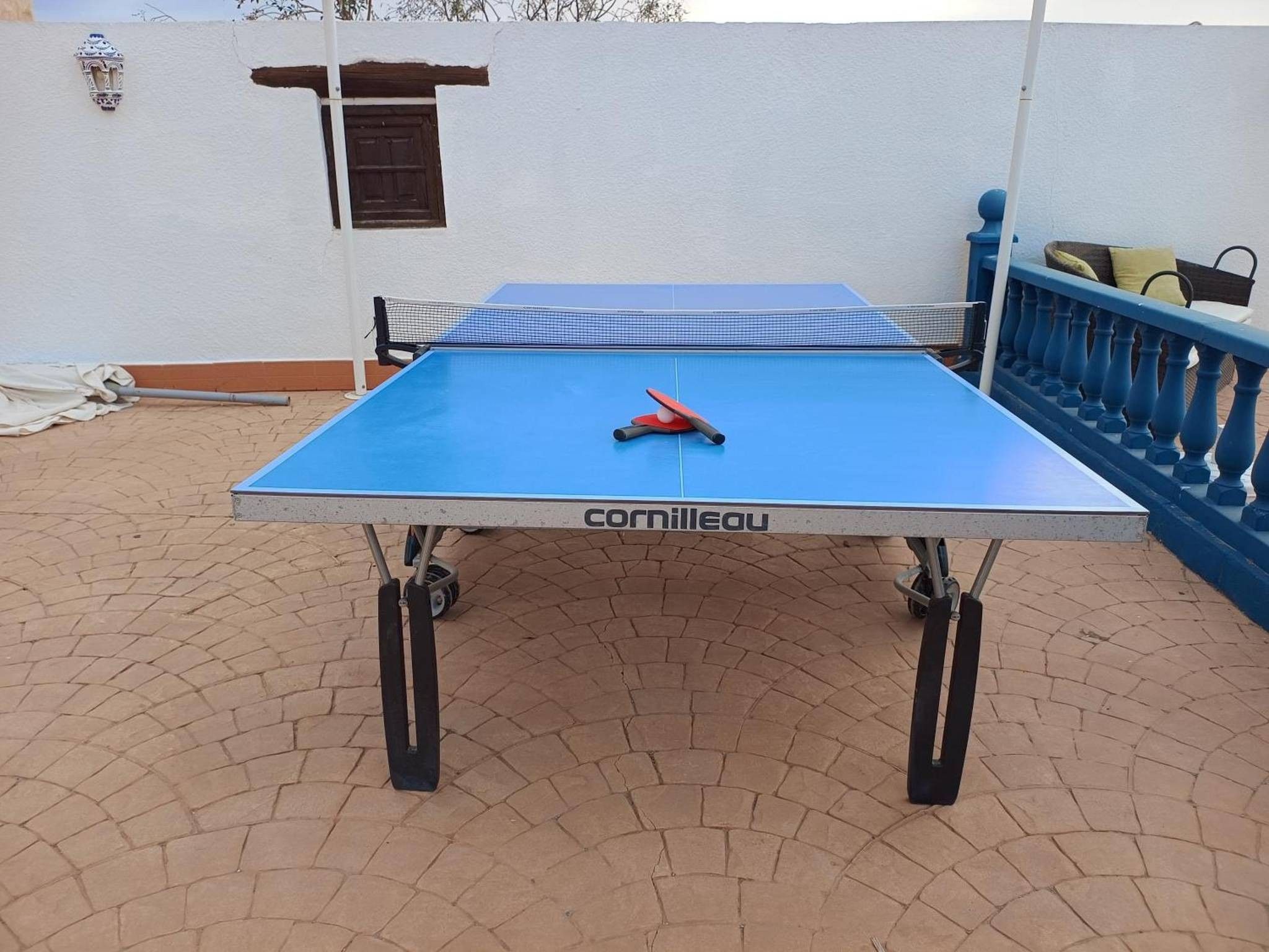 Una mesa de tenis de mesa con red, ubicada sobre una superficie pavimentada con una barandilla azul y una pared al fondo.