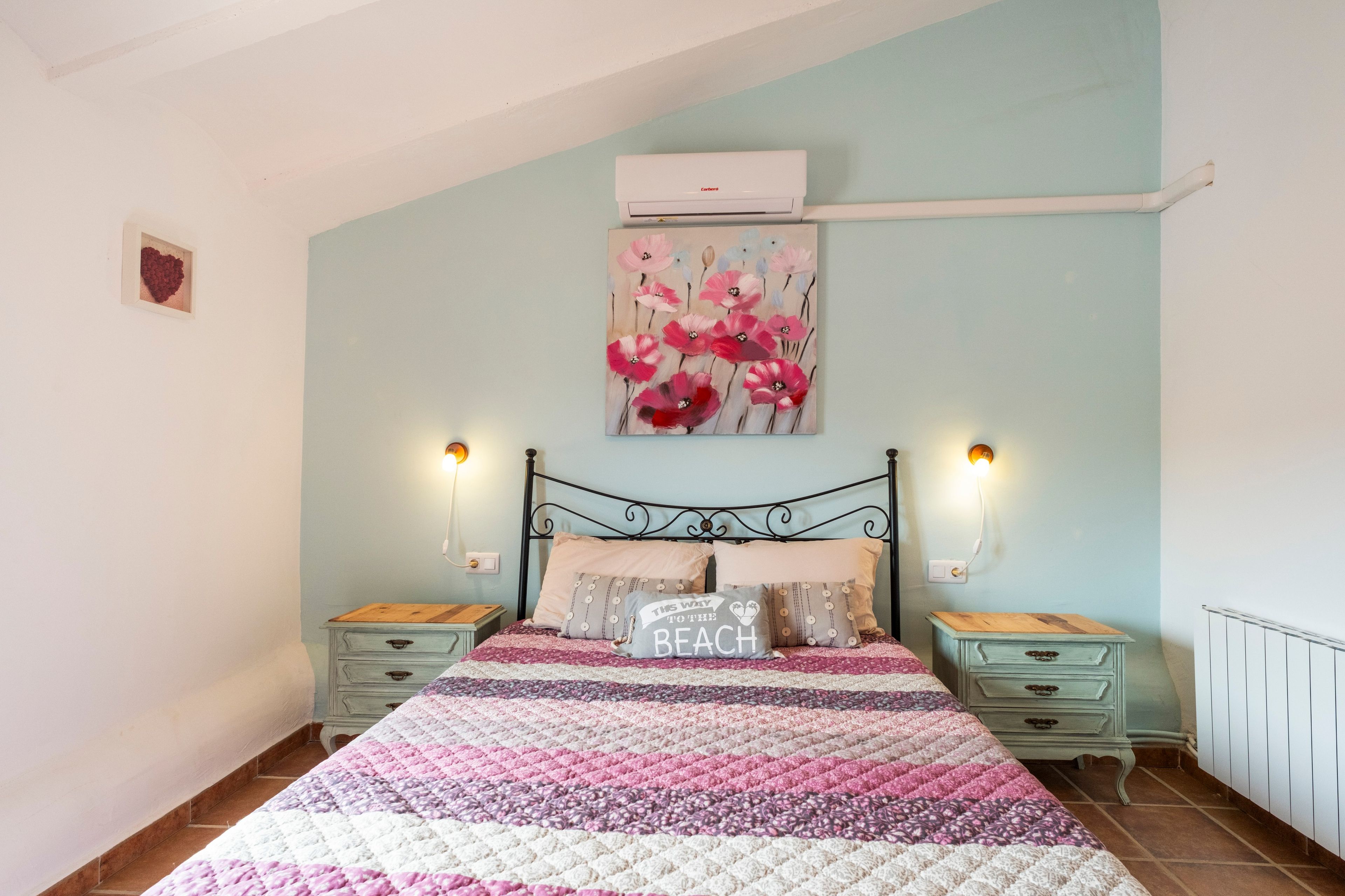 A bedroom with a bed, two bedside tables, a wall-mounted air conditioner, and a painting.