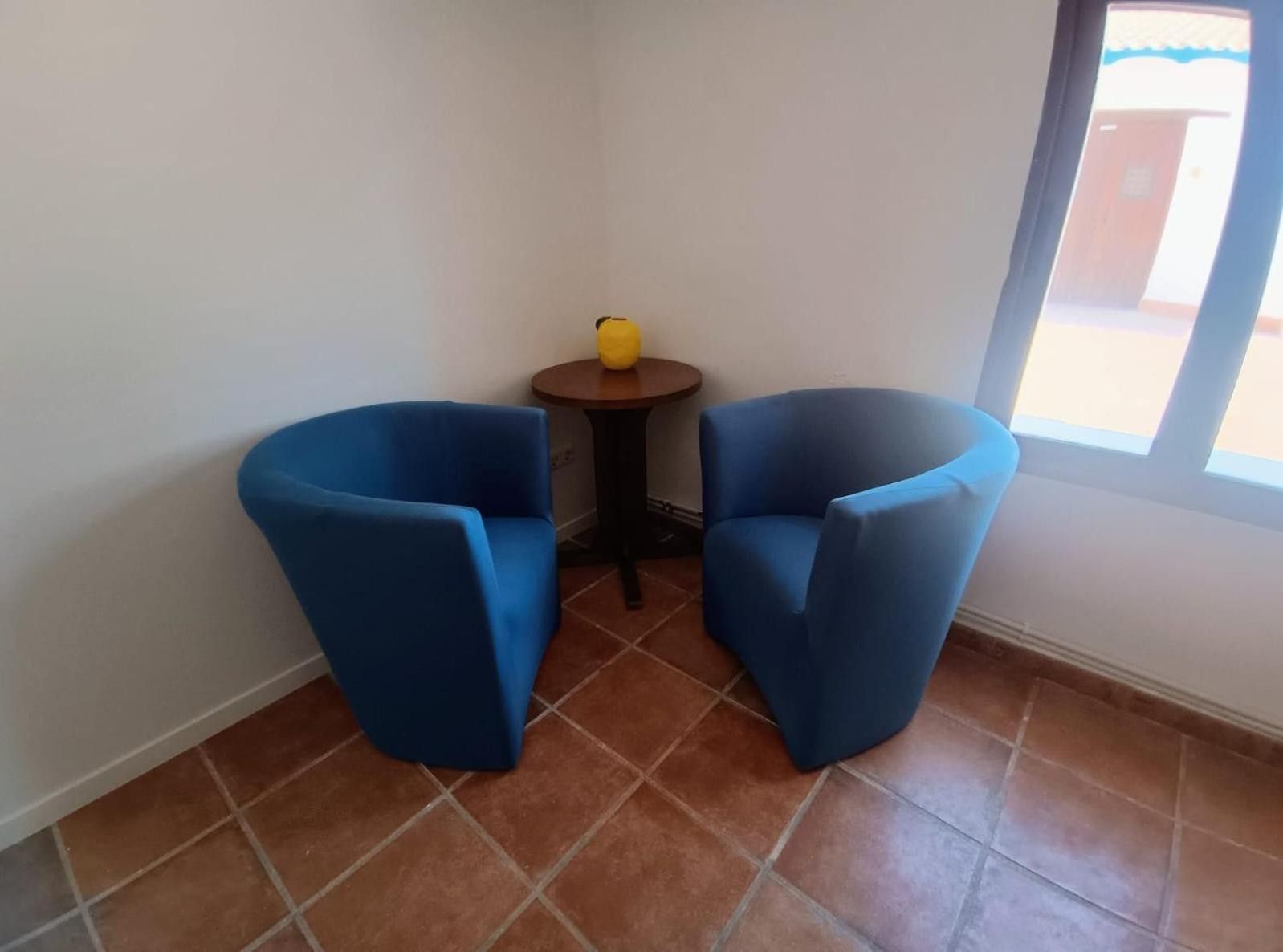 Une pièce avec deux fauteuils bleus, une petite table ronde sur laquelle se trouve un objet jaune, et une fenêtre donnant sur un paysage.