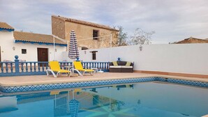 Une piscine dont les bords sont recouverts de carreaux bleus, des chaises longues jaunes et blanches, un parasol rayé et, en arrière-plan, un bâtiment blanc orné de motifs bleus.