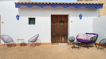 Un patio con un edificio blanco, una puerta de madera, dos sillas, una mesita y un sofá con cojines.
