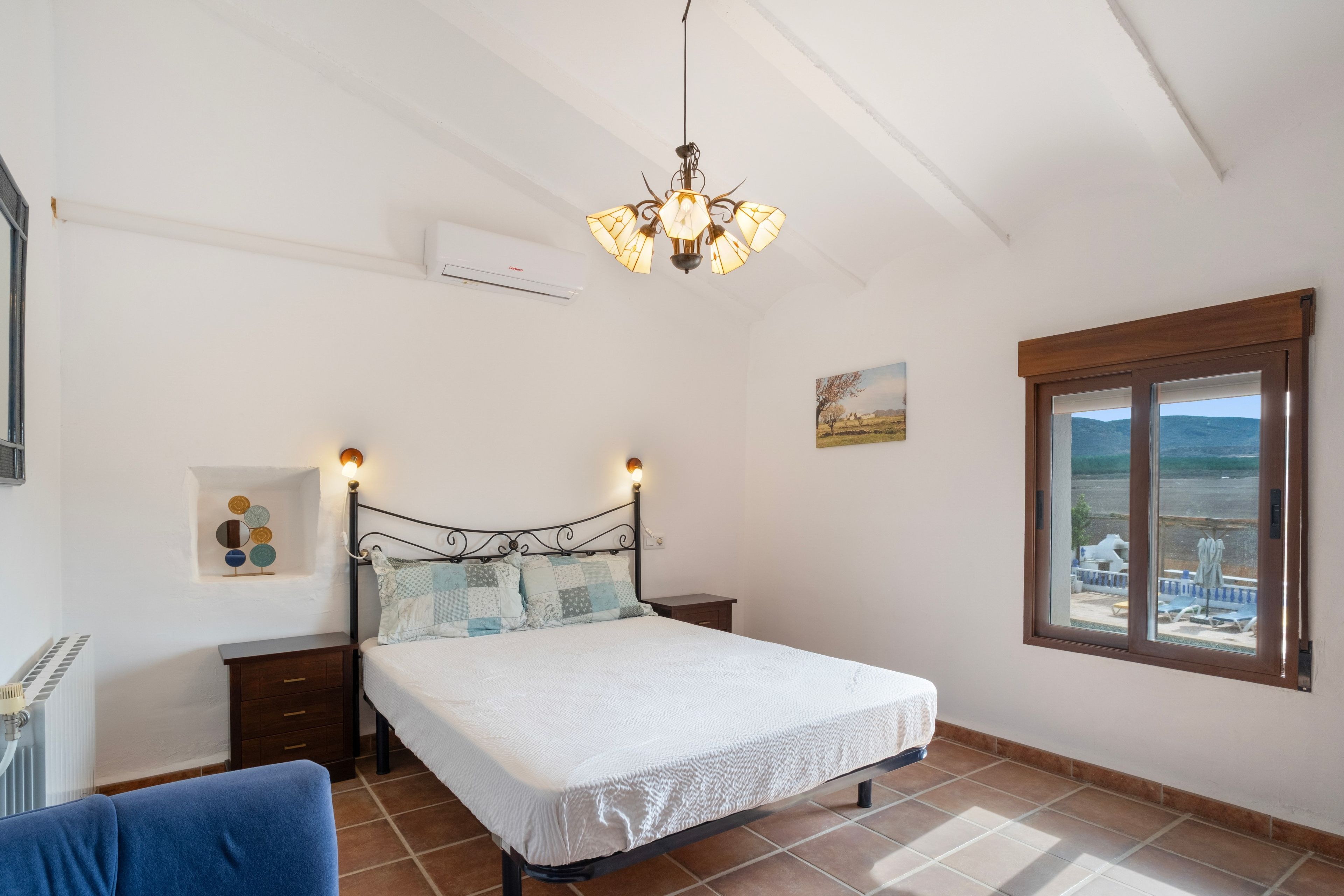 A bedroom with a bed, bedside tables, a radiator, a window with a view, and a chandelier.