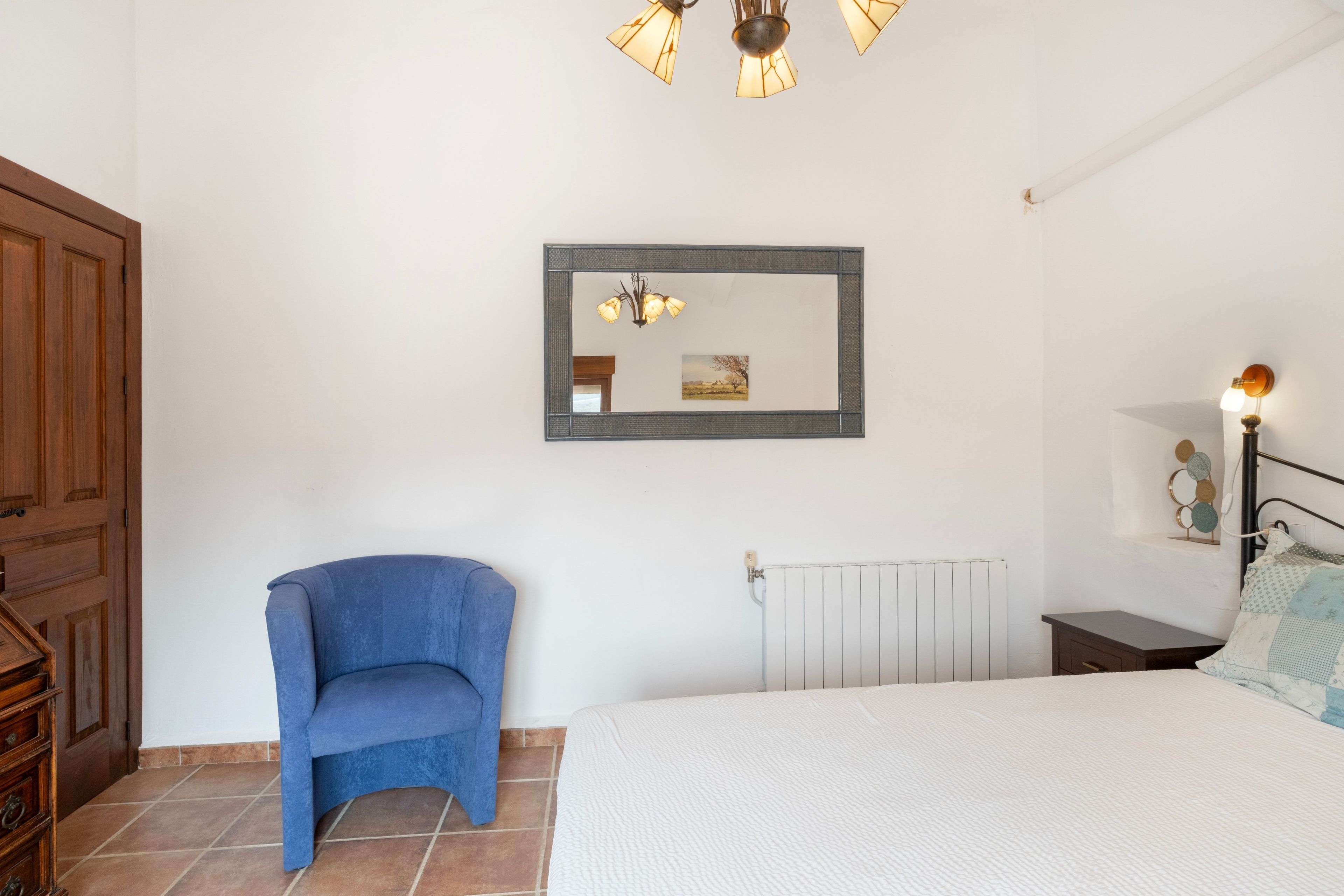 A bedroom with a bed, a chair, a radiator, a mirror, and a wall-mounted light fixture.