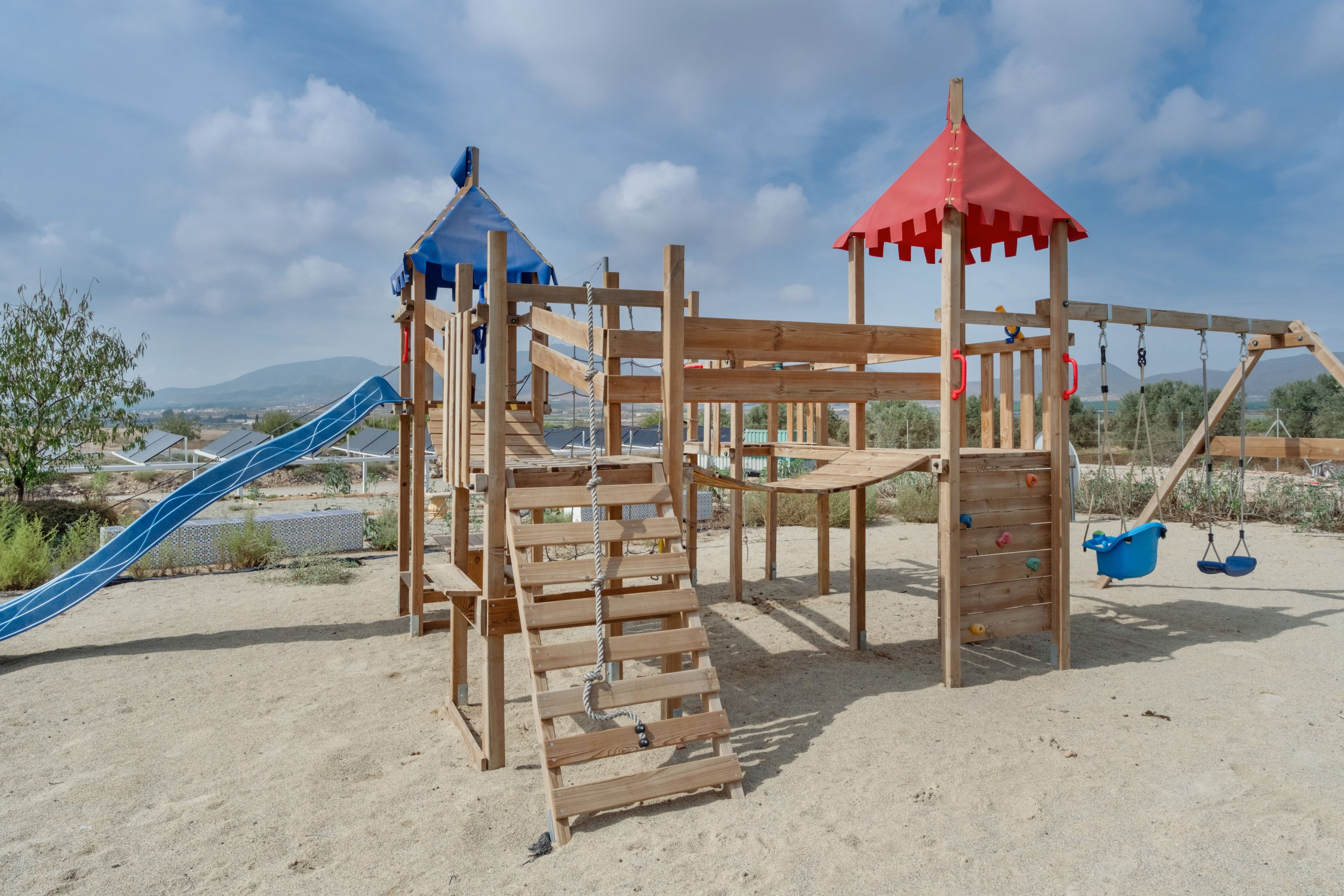 Un parque infantil de madera con tobogán y estructuras para escalar, situado sobre suelo arenoso bajo un cielo despejado.