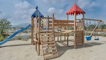 Un parque infantil de madera con tobogán y estructuras para escalar, situado sobre suelo arenoso bajo un cielo despejado.