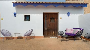 Une cour avec un bâtiment blanc, une porte en bois, deux chaises, une petite table et un canapé violet.