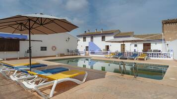 Zona junto a la piscina con sillones de descanso y sombrillas, cielo despejado y edificios tradicionales al fondo.