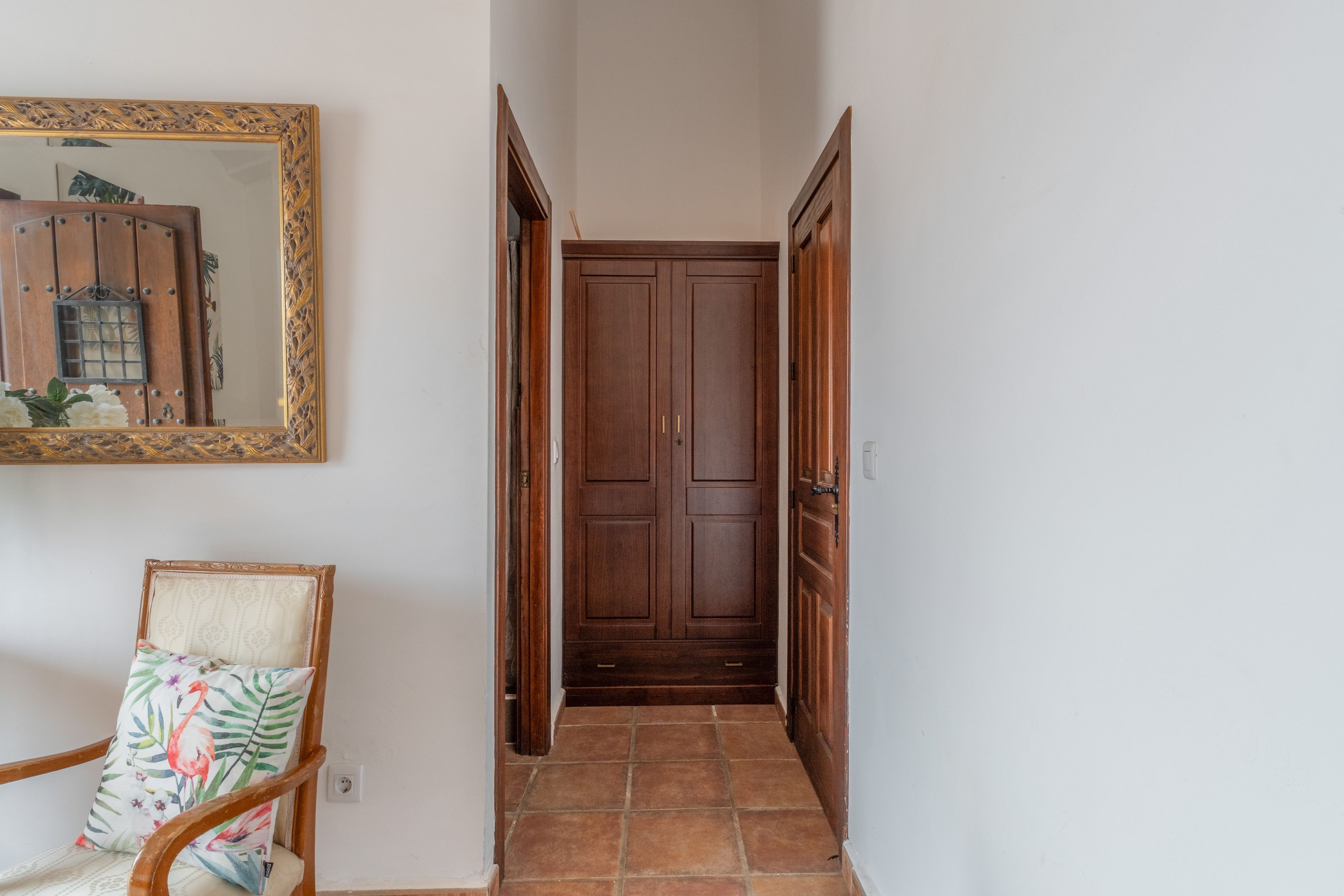 Un corridoio con una porta in legno, uno specchio con cornice floreale e una sedia con cuscino decorato con fiori.