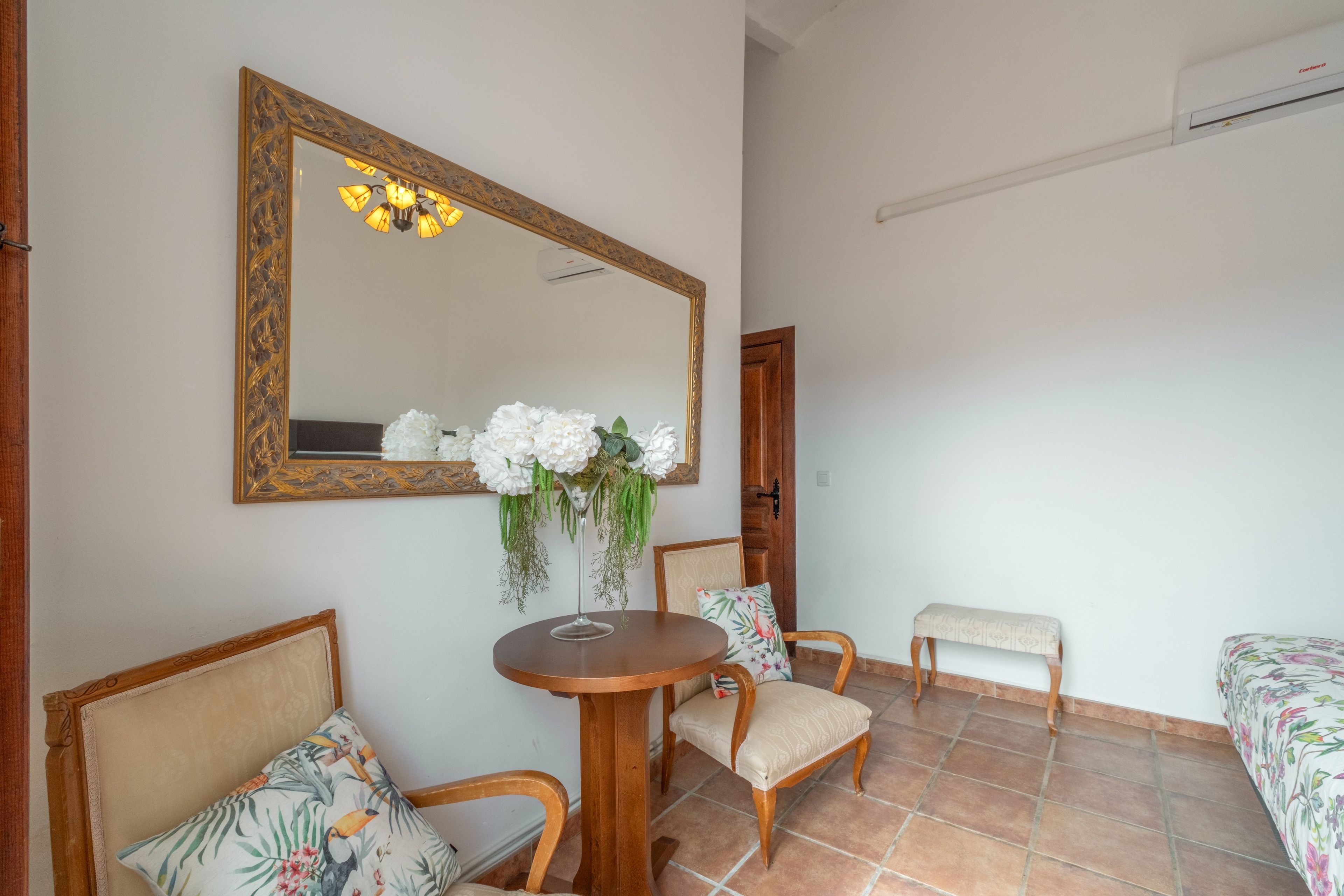 Ein Zimmer mit einem runden Holztisch, zwei Stühlen, einer Bank, einem Spiegel mit aufwendigem Rahmen und einer Blumenkomposition an der Wand.