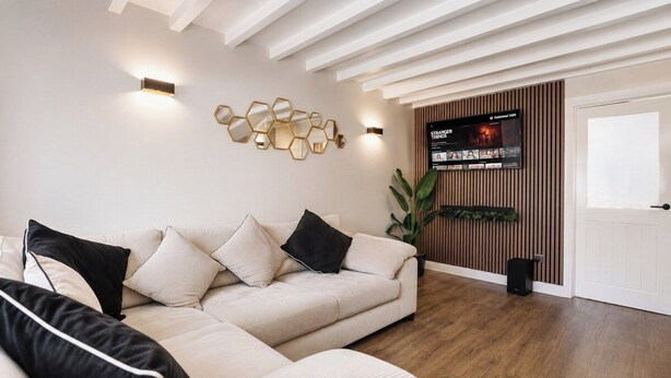 A modern living room with a beige sofa, black cushions, a wall-mounted TV, and a wooden floor. - Beautiful Abode in The Bath Cotswolds (Frome)