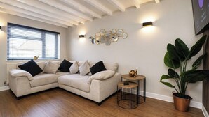 A living room with a beige sectional sofa, a small round table, a potted plant, and a wall-mounted light fixture. - Beautiful Abode in The Bath Cotswolds (Frome)