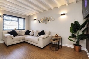 A living room with a beige sectional sofa, a small round table, a potted plant, and a wall-mounted light fixture. - Beautiful Abode in The Bath Cotswolds (Frome)
