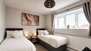 A bedroom with two beds, a bedside table with lamps, a radiator, and a window with curtains. - Beautiful Abode in The Bath Cotswolds (Frome)