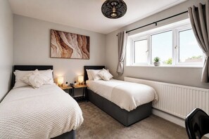 A bedroom with two beds, a bedside table with lamps, a radiator, and a window with curtains. - Beautiful Abode in The Bath Cotswolds (Frome)