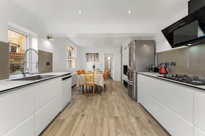 Una cocina moderna con armarios blancos, un comedor con mesa y sillas, y una pared de ladrillo visible a través de una ventana.