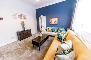 A living room with a dark wooden coffee table, a beige sofa with patterned cushions, and a blue accent wall. - The Sunderland Haven | 6BR Home for 14 (Sunderland)