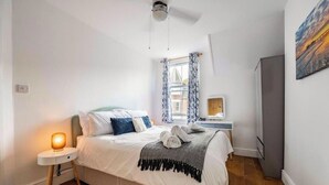 A bedroom with a bed, a bedside lamp, a window with curtains, a ceiling fan, and a painting on the wall. - Modern Cromer Coastal Flat (Norfolk)