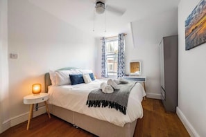 Um quarto com cama, abajur, janela com cortinas, ventilador de teto e um quadro na parede.