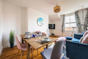 Uma sala de jantar com uma mesa de madeira posta para quatro pessoas, uma planta em vaso e um sofá azul ao fundo.