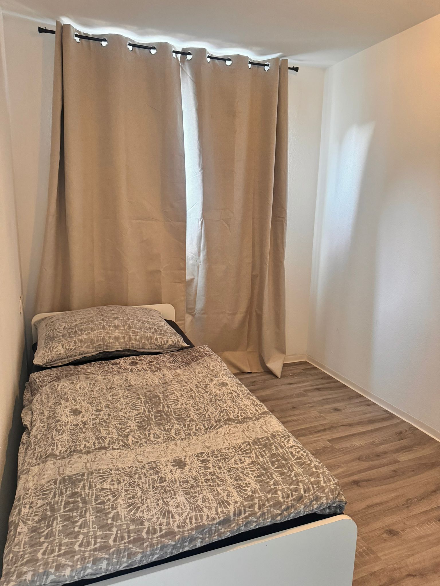 A single bed with a patterned bedspread and two pillows, a wooden floor, and a curtain with a rod.