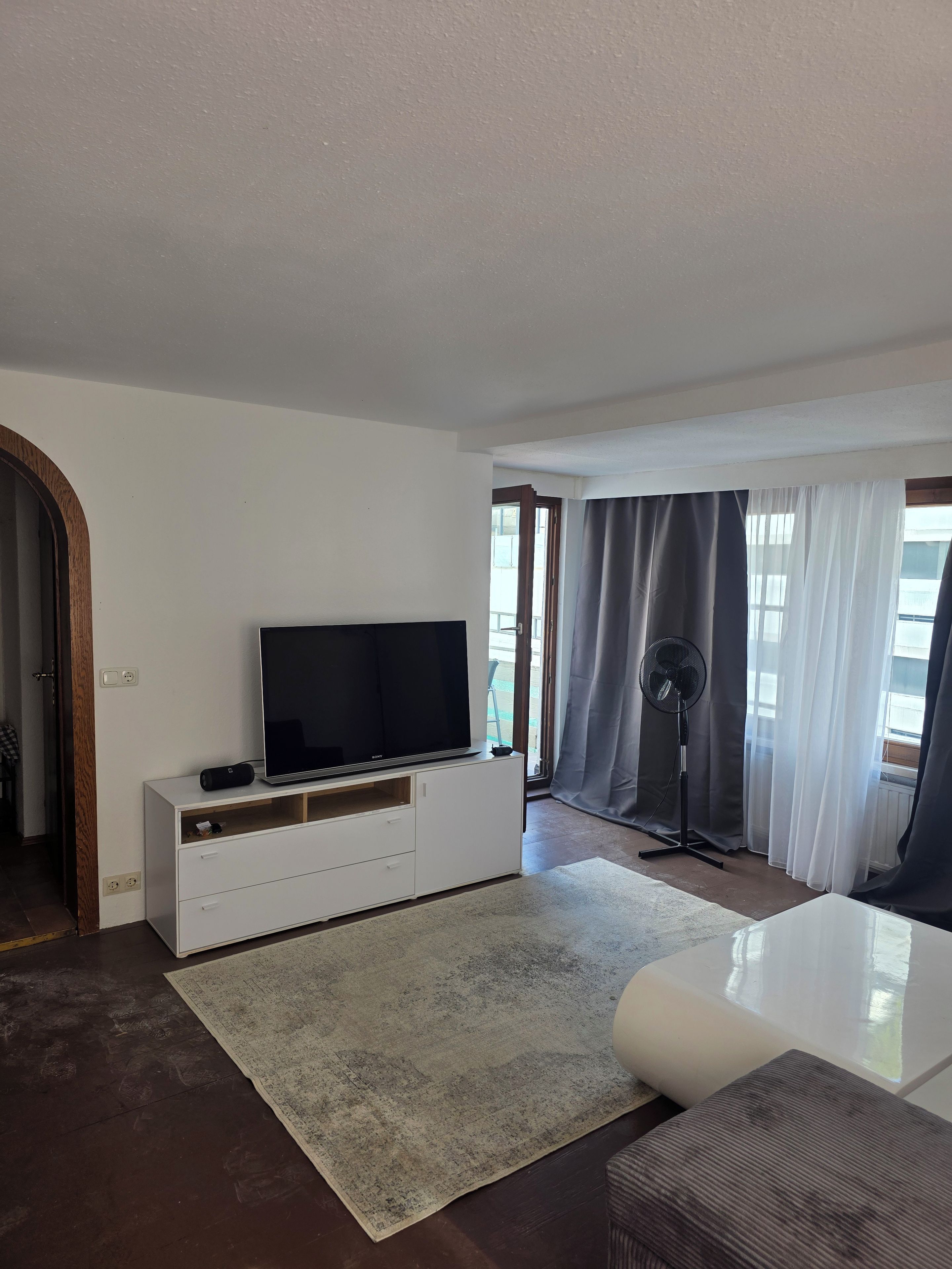 Een moderne woonkamer met een flatscreen televisie op een witte kast, een staande ventilator en een groot raam met gordijnen.
