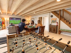 A room with a foosball table, a television displaying a sports game, and a staircase. - Beach Days, Game Nights Near Downtown Jensen Beach (Jensen Beach)