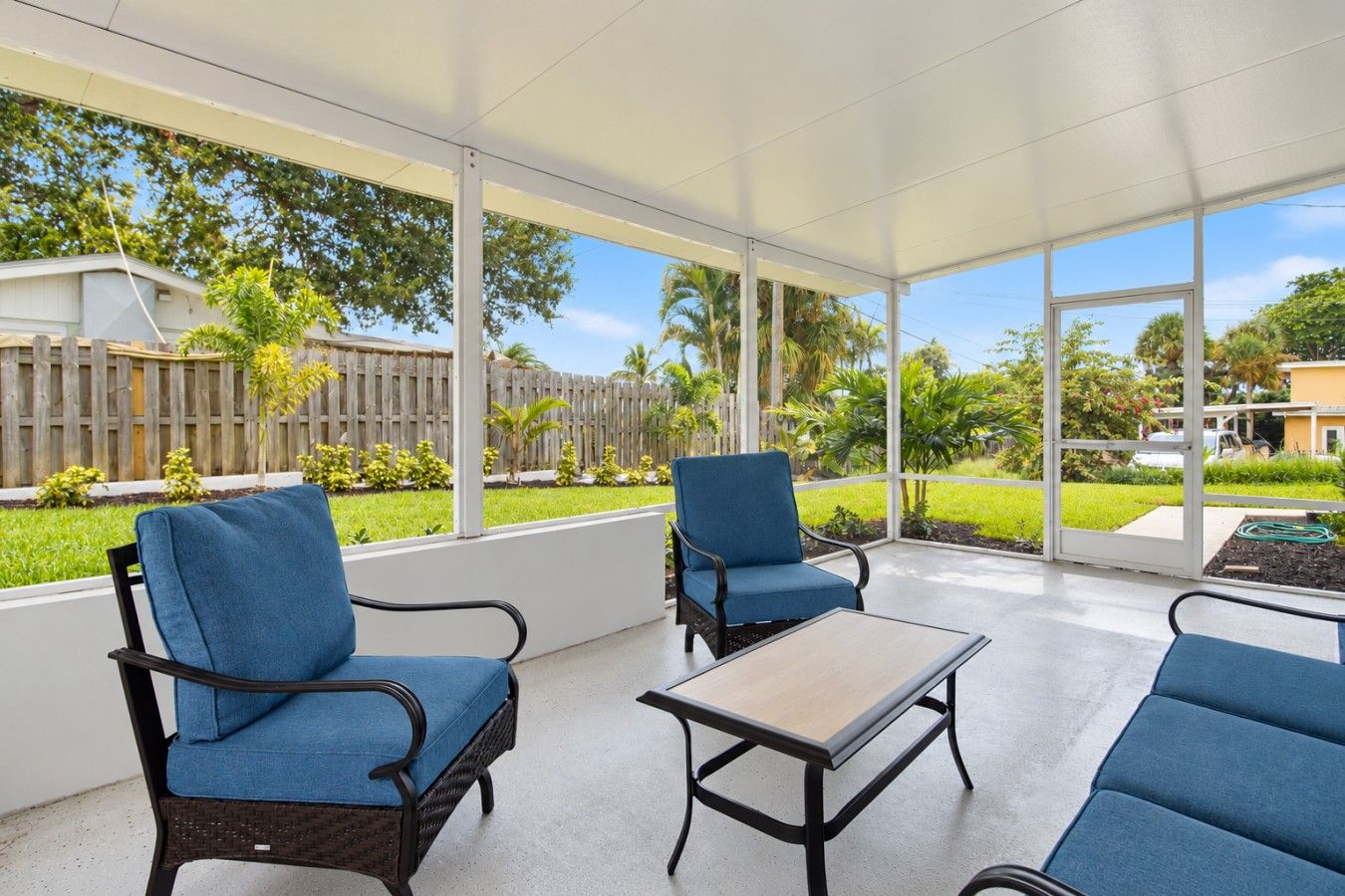 A covered patio with two blue cushioned chairs and a small table, overlooking a well-maintained garden and neighboring houses.