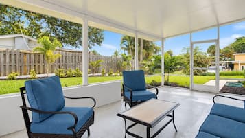 A covered patio with two blue cushioned chairs and a small table, overlooking a well-maintained garden and neighboring houses.