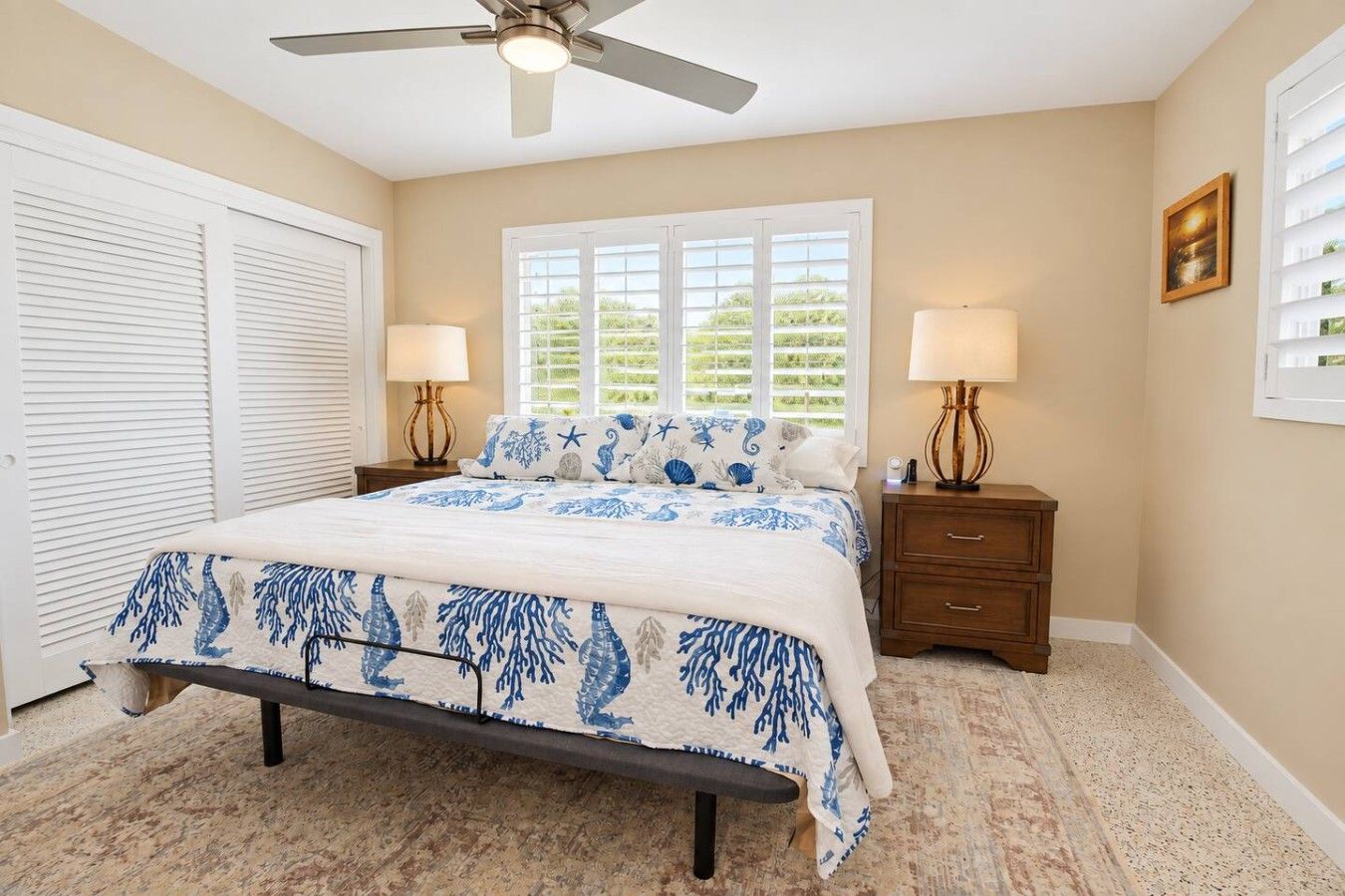 A bedroom with a bed, two bedside tables with lamps, a ceiling fan, a window with blinds, and a door with a shutter.
