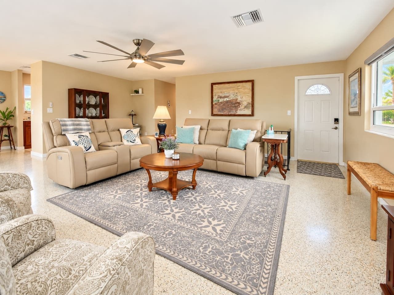 A spacious living room with a beige sofa set, a coffee table, patterned rugs, and a ceiling fan.