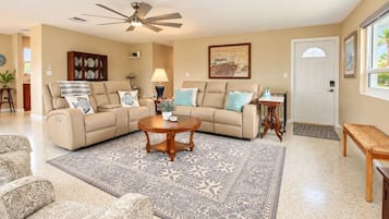 A spacious living room with a beige sofa set, a coffee table, patterned rugs, and a ceiling fan.