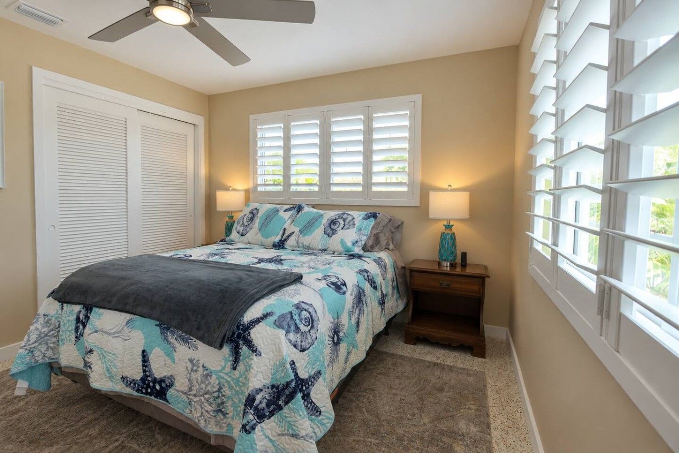 A bedroom with a bed, a ceiling fan, a window with blinds, and a nightstand with a lamp.