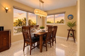 Uma área de jantar com uma mesa posta para seis pessoas, rodeada por cadeiras, e vista para palmeiras através de grandes janelas.