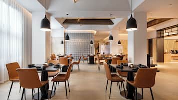 A modern dining area with multiple tables and chairs, a high ceiling with exposed beams, and pendant lighting.