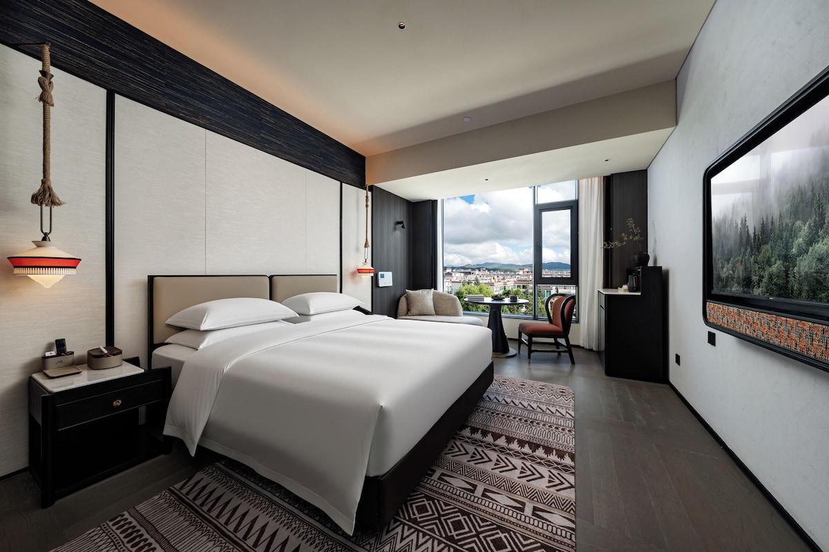A modern hotel room with a large bed, a desk, a chair, and a view of the city through large windows.