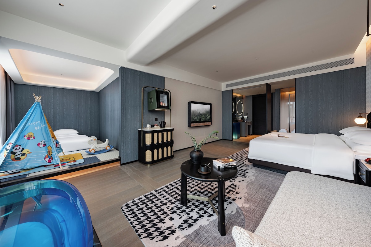 A modern hotel room with a bed, a desk, a chair, a small table with a vase, and a blue play tent.