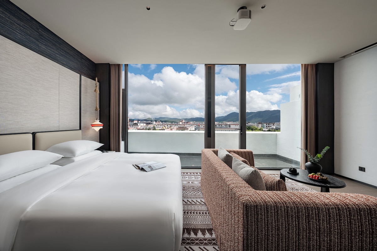A modern hotel room with a large bed, a sofa, a small table with a plant, and a view of a cityscape through large windows.